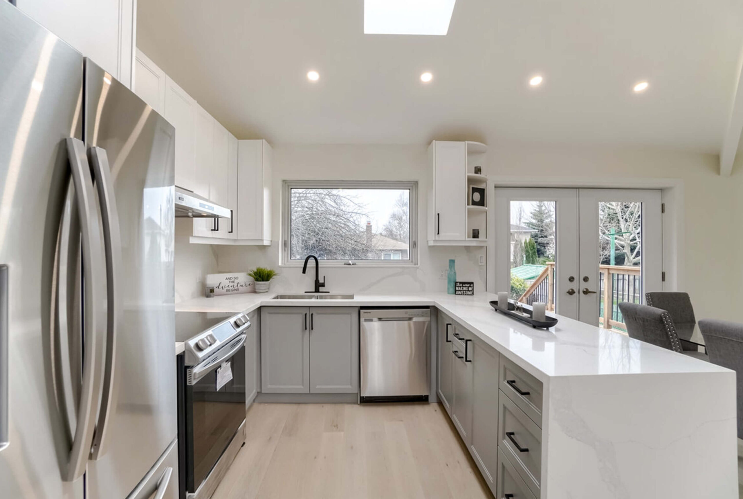 Lifestyle Construction Modern kitchen with stainless steel appliances, white countertops, gray cabinets, and a window above the sink. French doors lead to a deck, and recessed lighting is installed in the ceiling. This Toronto Renovation by Lifestyle Construction combines functionality with contemporary elegance. Home Renovation In The GTA