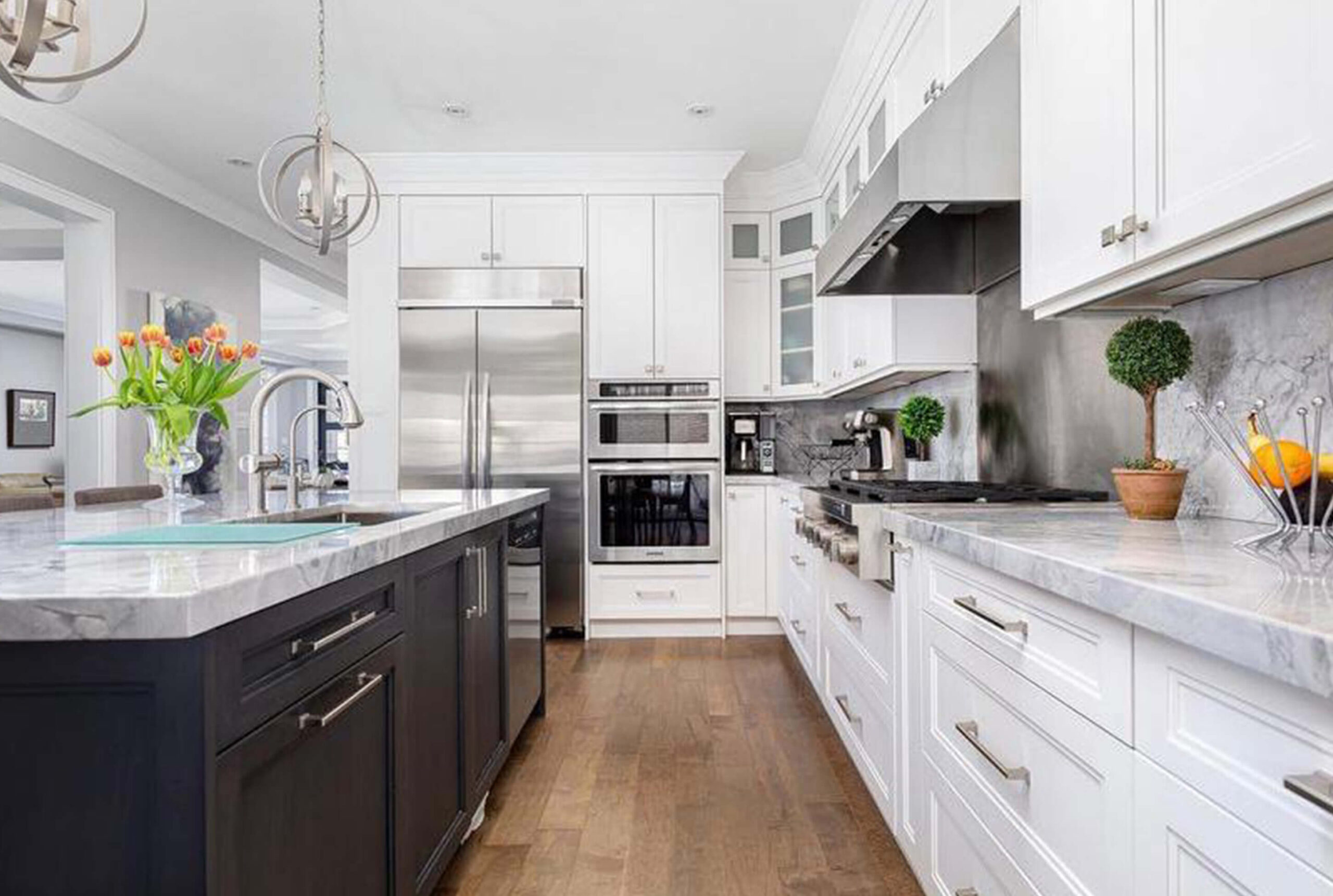 Lifestyle Construction A modern kitchen with white cabinetry, stainless steel appliances, and wooden flooring. The large island features a sink, a vase of tulips, and a bowl of oranges. Perfectly crafted by Lifestyle Construction during a Home Renovation Vaughan project. Home Renovation In The GTA