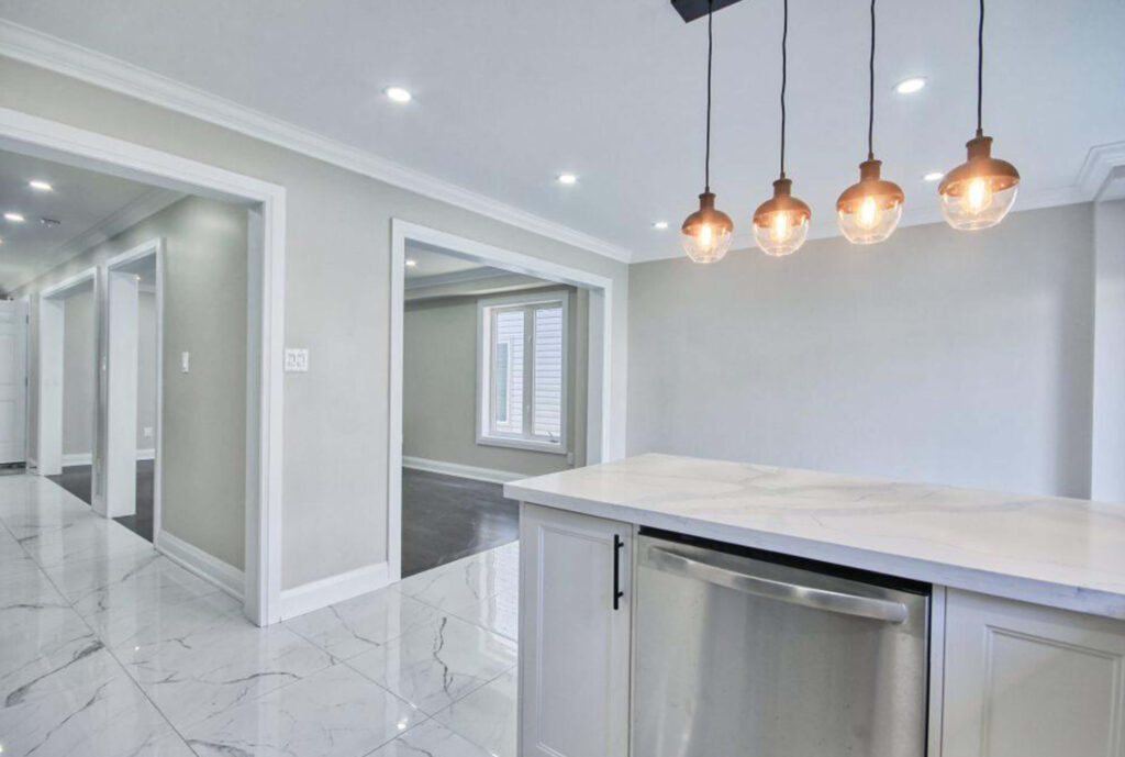 Lifestyle Construction Modern kitchen with marble flooring, white countertops, stainless steel dishwasher, and pendant lighting. The adjacent hallway leads to additional rooms with large windows and similar flooring, all part of a Toronto Renovation project aimed at enhancing style and comfort. Home Renovation In The GTA