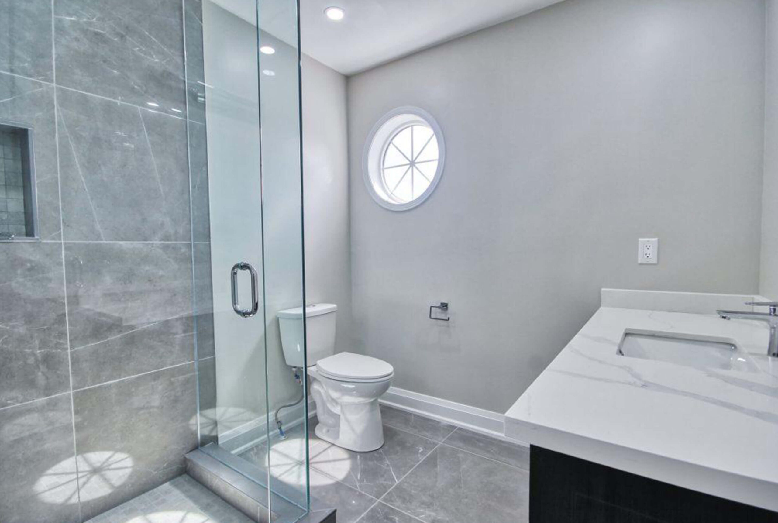 Lifestyle Construction Modern bathroom featuring a glass-enclosed shower, a round window, a toilet, and a white marble sink vanity over a glossy tile floor. Perfect for those seeking sophisticated elegance in their home renovation Vaughan. Home Renovation In The GTA