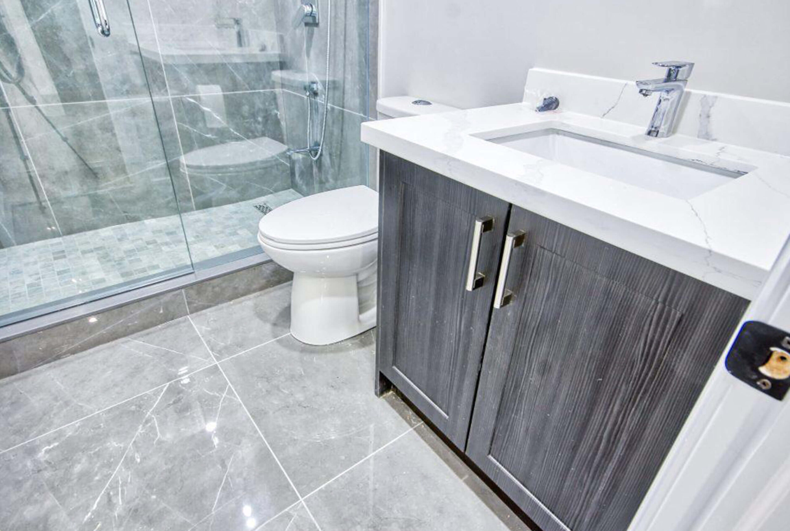 Lifestyle Construction A modern bathroom by Lifestyle Construction featuring a glass shower enclosure, a toilet, and a vanity with a white countertop and dark wood cabinetry on a polished tile floor. This Toronto renovation showcases sleek design and meticulous craftsmanship. Home Renovation In The GTA