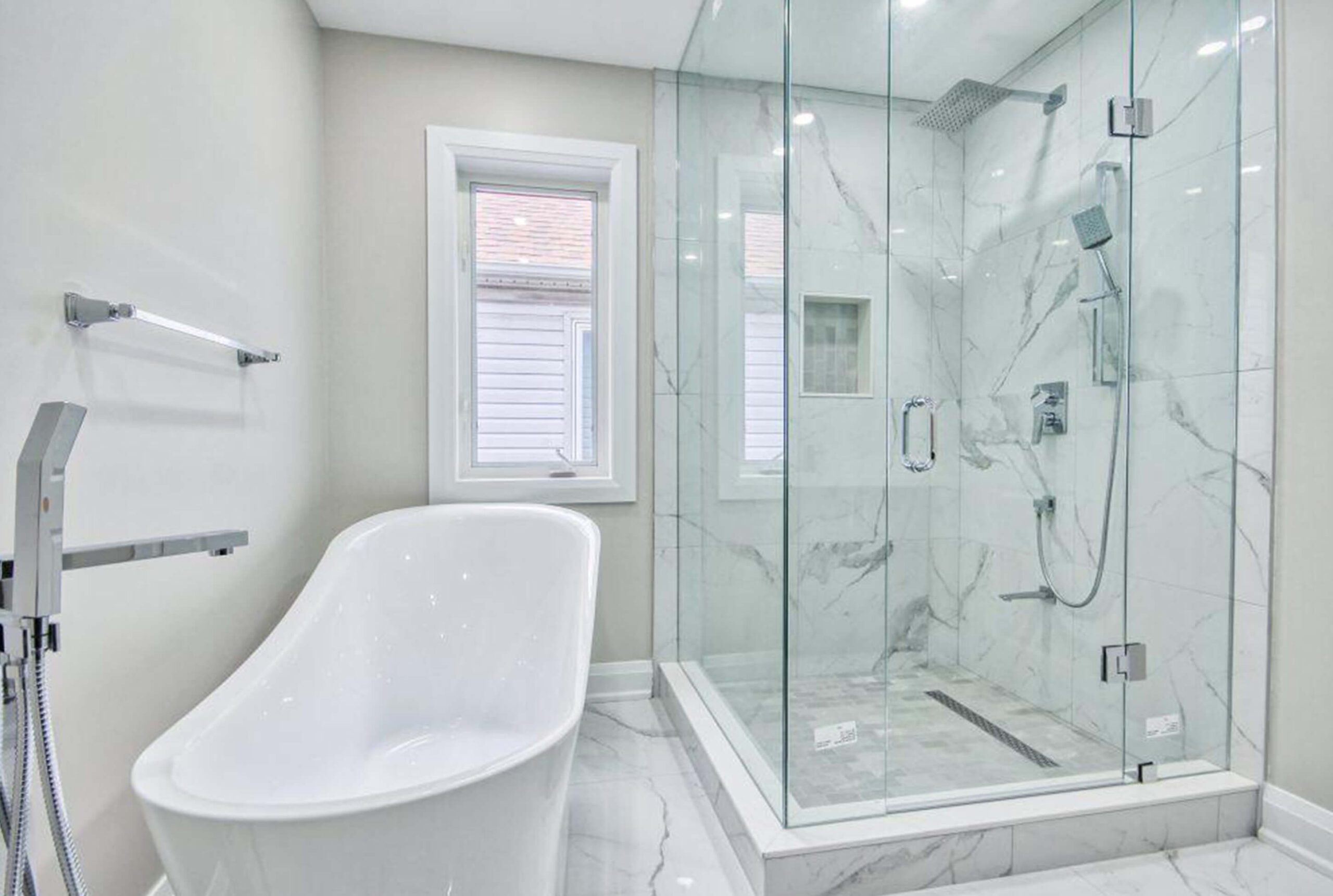 Lifestyle Construction A modern bathroom by Lifestyle Construction features a freestanding white bathtub, glass-enclosed shower area, and a small window. The walls and floor are tiled with a light gray marble pattern, showcasing the elegance of Toronto Renovation expertise. Home Renovation In The GTA