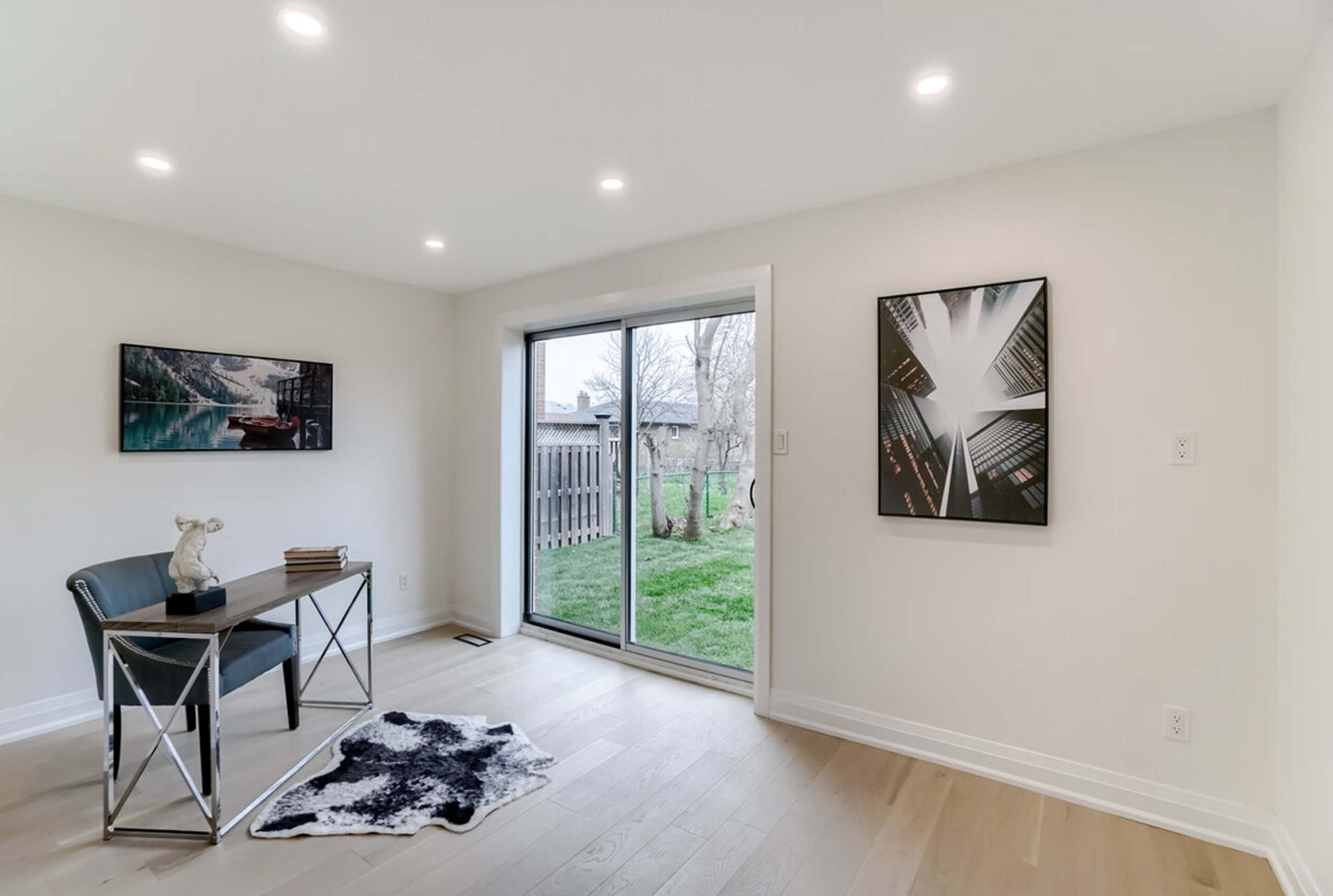 Lifestyle Construction Minimalist room with a desk, chair, and decorative items. Large glass door opens to a backyard. Two modern art pieces on white walls. Light wooden flooring with a small cowhide rug—a prime example of Toronto Renovation finesse. Home Renovation In The GTA