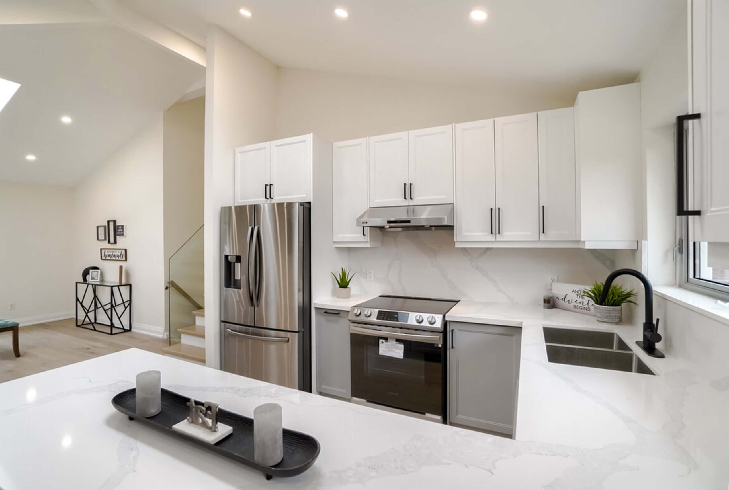 Lifestyle Construction Modern kitchen with white cabinets, stainless steel appliances, marble countertops, and a black faucet on a white sink. A tray with candles is on the island, and a staircase is visible in the background. Perfect for those embracing a Home Renovation Vaughan lifestyle. Home Renovation In The GTA