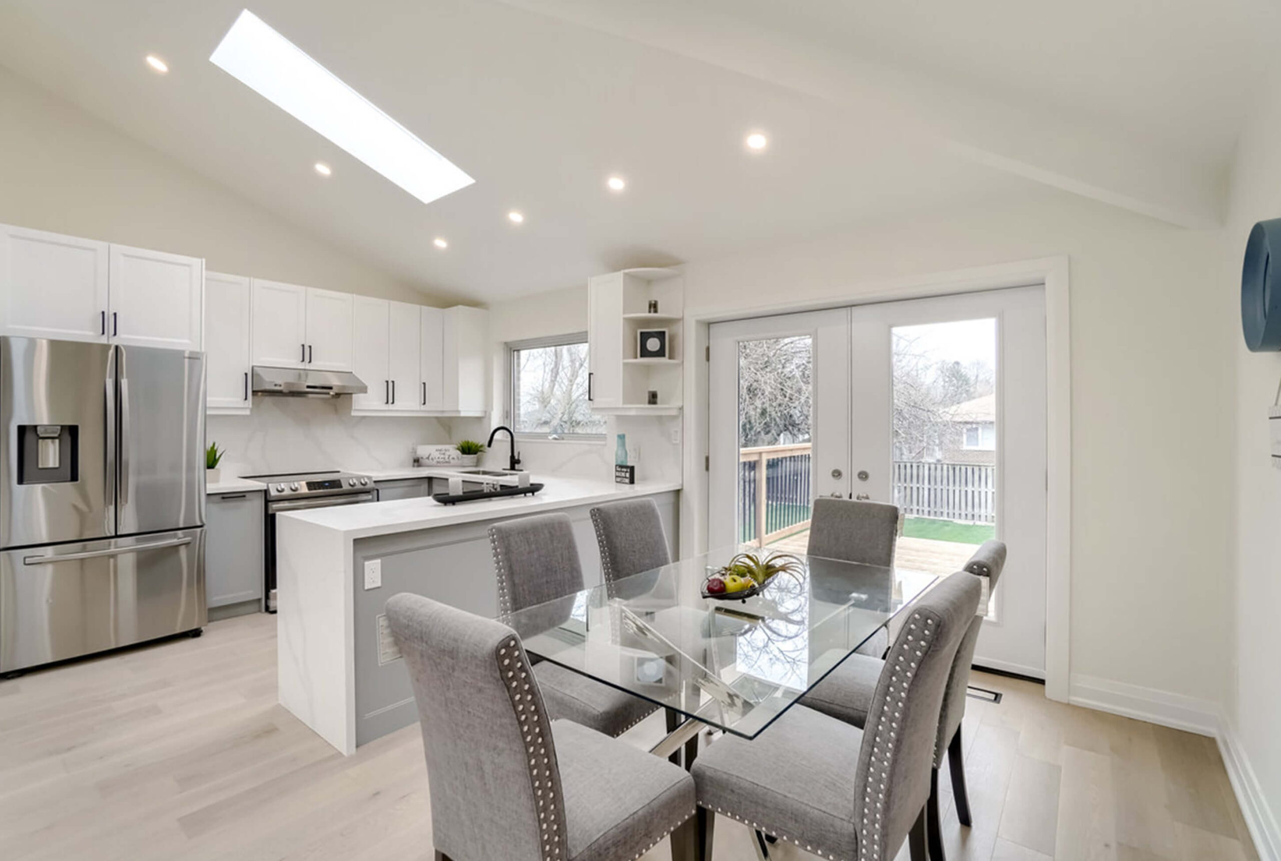 Lifestyle Construction Modern kitchen with white cabinets, stainless steel appliances, a central island, and glass dining table. The sliding glass doors open to a backyard, reflecting the elegance of Toronto Renovation style for a contemporary home. Home Renovation In The GTA