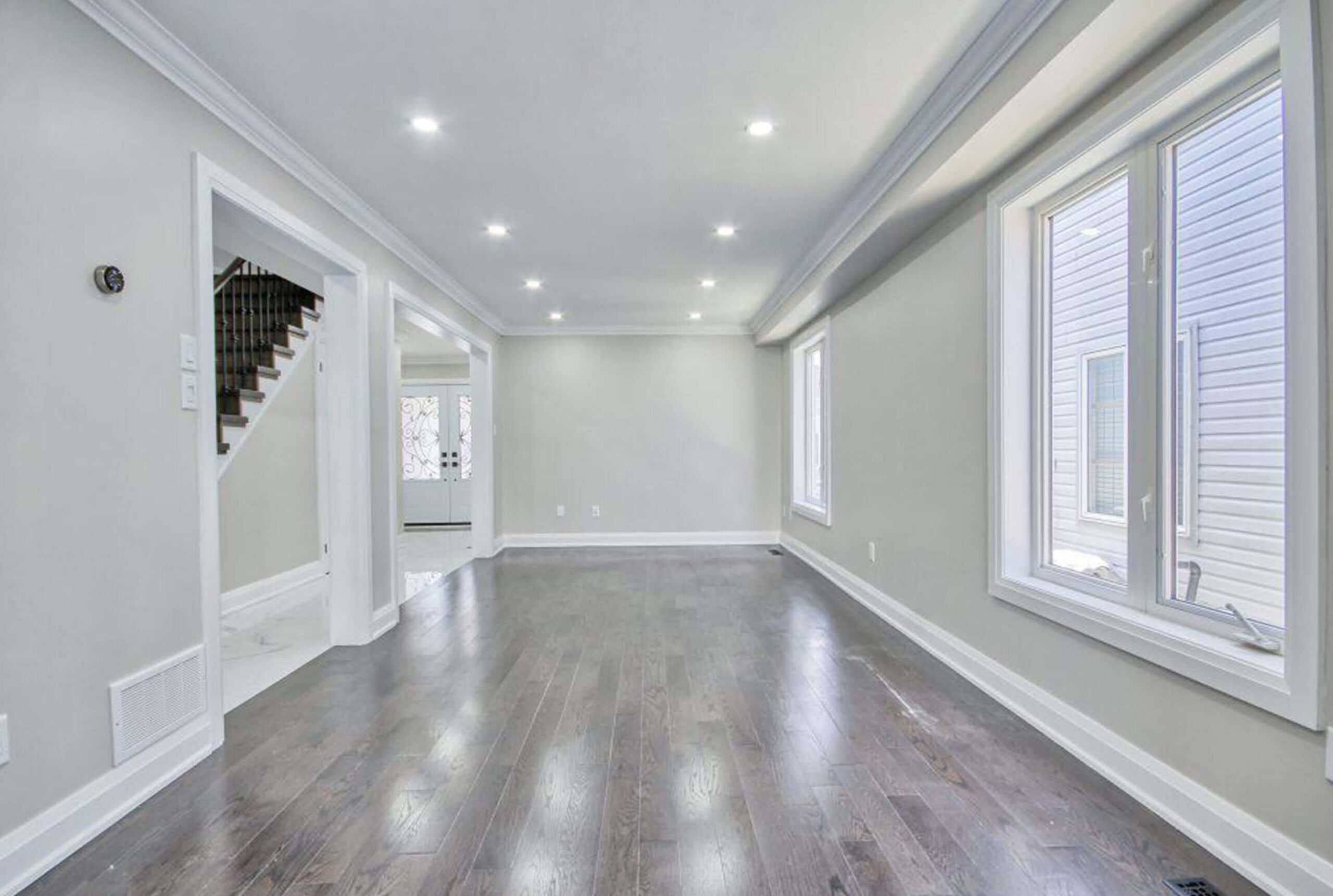 Lifestyle Construction A bright, empty living room with hardwood floors, recessed lighting, large windows, and white walls. An open doorway leads to a staircase and another area of the house. Perfect for those seeking Lifestyle Construction as part of their Toronto Renovation dreams. Home Renovation In The GTA