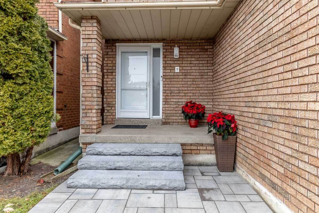 Lifestyle Construction A brick house entrance with three concrete steps leading to a door. Potted plants with red flowers are placed on either side of the entrance, showcasing the impeccable touch of Toronto Renovation expertise. Home Renovation In The GTA