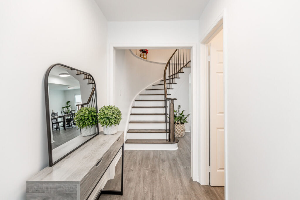 Lifestyle Construction A hallway featuring a wooden console table and a mirror on the left. Ahead, a curved staircase with a metal railing adds elegance. The wood flooring complements the minimal decor, including two potted plants. Ideal for those seeking Toronto Renovation inspiration or Home Renovation Vaughan projects. Home Renovation In The GTA