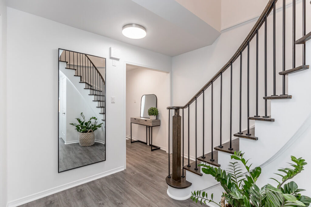 Lifestyle Construction A modern hallway in Vaughan features a large mirror, a small console table, potted plants, and a curved staircase with sleek black railings. This stylish space embodies the essence of Toronto renovation trends while highlighting contemporary lifestyle construction. Home Renovation In The GTA