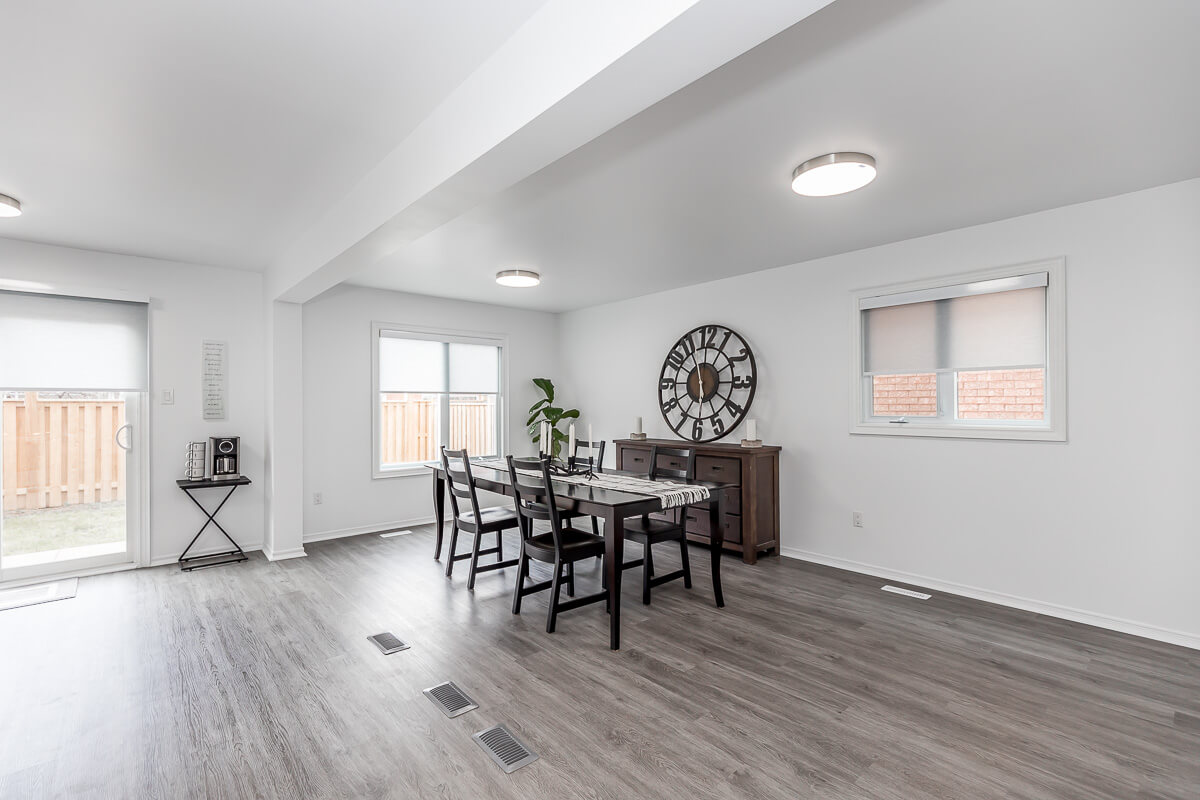 Lifestyle Construction A modern dining room with a large clock on the wall, a dining table for six, a sideboard, and two windows providing natural light. The grey laminate floor and white-painted walls showcase the elegance of Toronto Renovation expertise. Home Renovation In The GTA