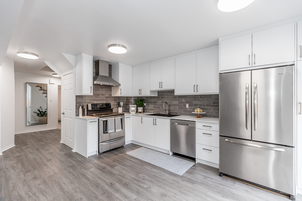 Lifestyle Construction A modern kitchen with white cabinets, stainless steel appliances including a fridge, oven, and dishwasher, gray backsplash tiles, and gray wood flooring. A mat is placed in front of the sink. This space exemplifies Toronto Renovation excellence in home renovation Vaughan projects. Home Renovation In The GTA