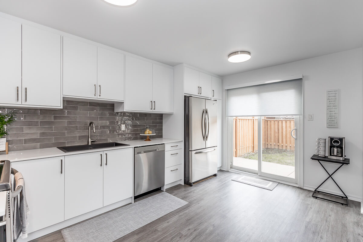 Lifestyle Construction Modern kitchen with white cabinets, gray tile backsplash, stainless steel appliances, and sliding glass doors leading to a fenced yard—a perfect example of excellent Home Renovation in Vaughan. Home Renovation In The GTA