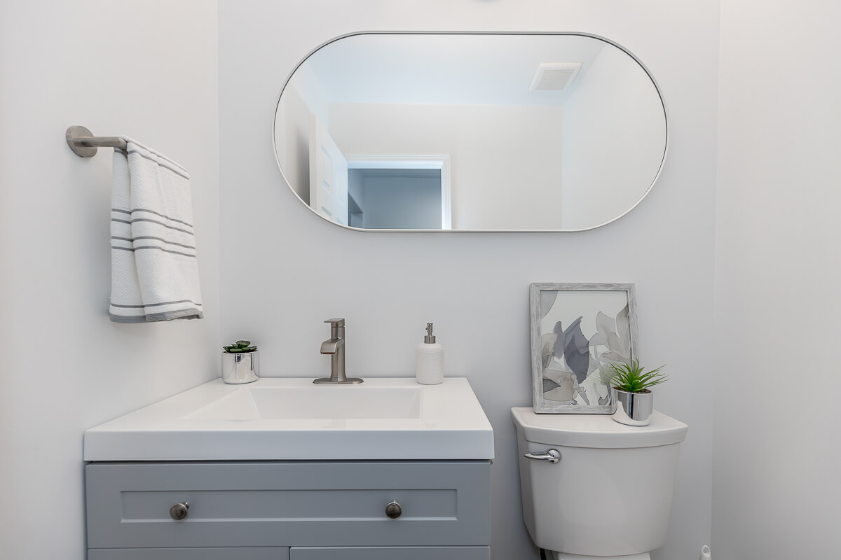 Lifestyle Construction Modern bathroom with a rectangular mirror, light gray vanity, silver faucet, hand towel, potted plants, and wall art above the toilet. Perfect for those undertaking a Toronto Renovation or seeking inspiration from Home Renovation Vaughan projects. Home Renovation In The GTA