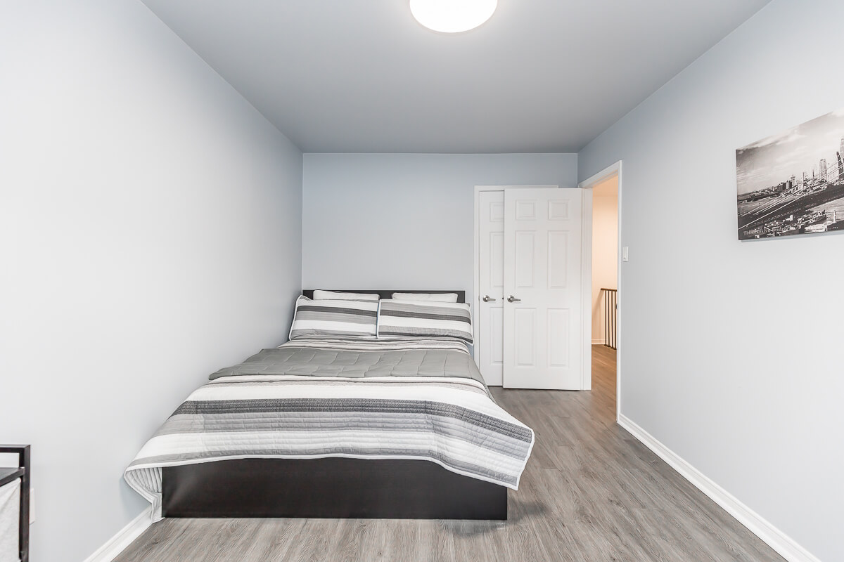 Lifestyle Construction A small bedroom with light gray walls, a bed with gray and white striped bedding, a closed door, a black and white framed picture on the wall, and wood-look flooring showcases the attention to detail characteristic of Toronto Renovation projects. Home Renovation In The GTA