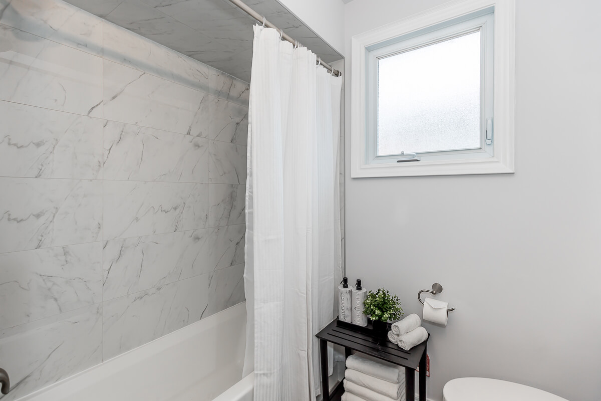 Lifestyle Construction A clean bathroom features a white tiled bathtub with a shower curtain, a window, and a black side table holding toiletries, towels, and a small plant—a perfect example of elegant design by Home Renovation Vaughan. Home Renovation In The GTA