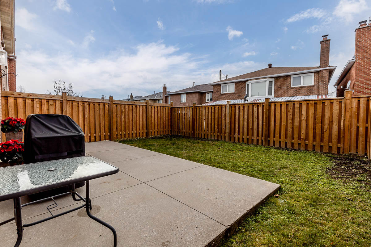 Lifestyle Construction A fenced backyard with a concrete patio, a glass table with black legs, and a covered grill. The yard is partially grassed and surrounded by a wooden fence, hinting at the superior Lifestyle Construction seen in Toronto renovations. Houses are visible beyond the fence. Home Renovation In The GTA