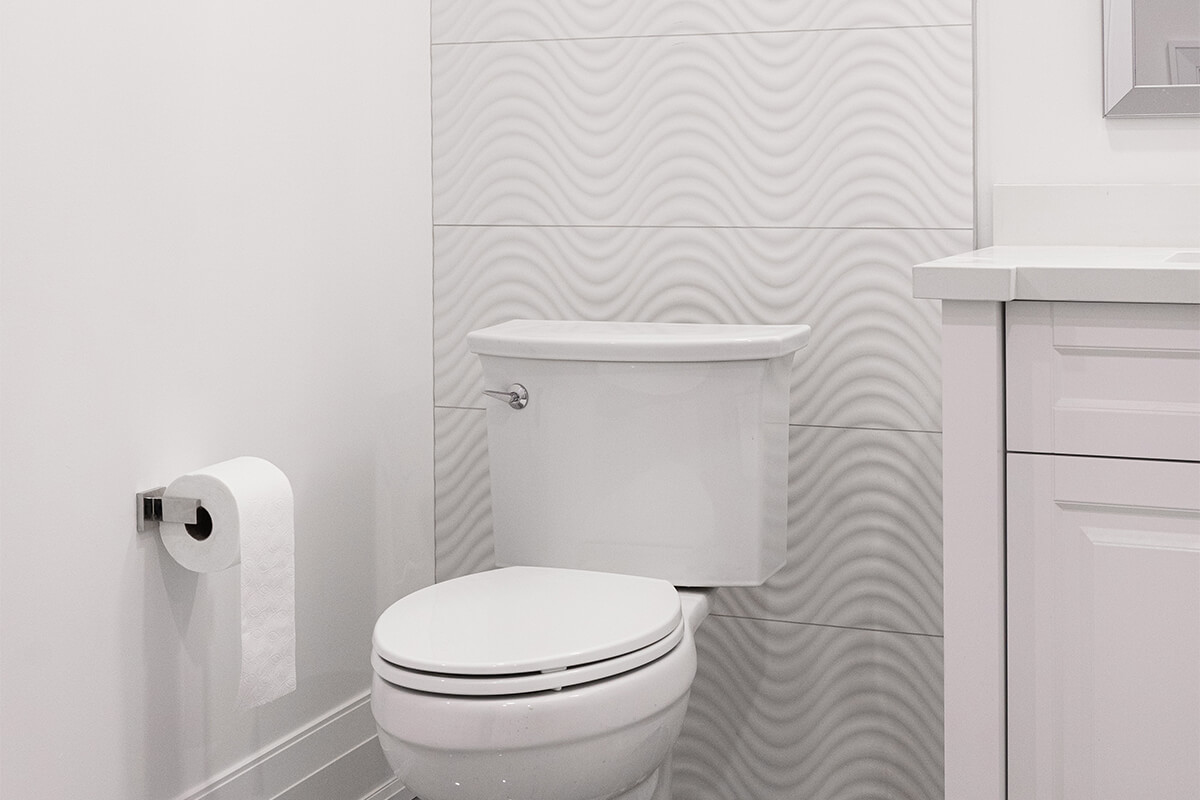 Lifestyle Construction A modern white bathroom by Lifestyle Construction featuring a toilet, textured wave-patterned wall tiles behind it, a roll of toilet paper on the left, and a white vanity with countertop on the right, showcasing high-quality craftsmanship from a Toronto Renovation. Home Renovation In The GTA