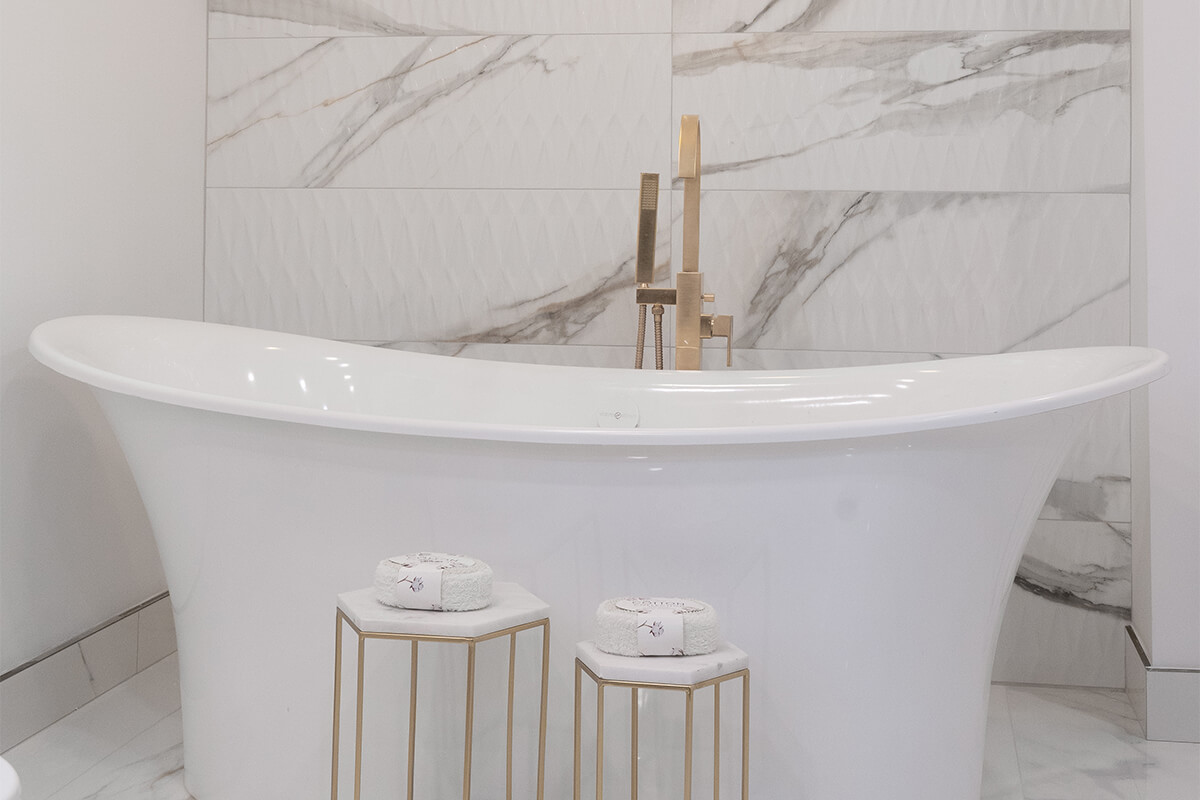 Lifestyle Construction A modern bathroom features a sleek white freestanding tub with tall, gold-tone fixtures, two small gold stools with folded white towels, and a marbled tile wall background, all expertly crafted by Home Renovation Vaughan. Home Renovation In The GTA