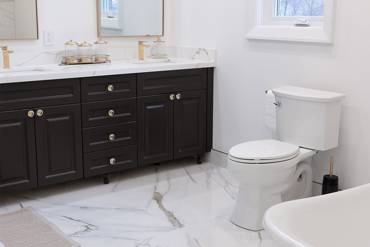 Lifestyle Construction A modern bathroom in a Toronto Renovation with white marble floors, a white toilet, and a dark wood vanity featuring dual sinks. The vanity boasts gold hardware and a marble countertop, perfectly blending style and functionality. A beautifully designed tub completes this Lifestyle Construction project. Home Renovation In The GTA