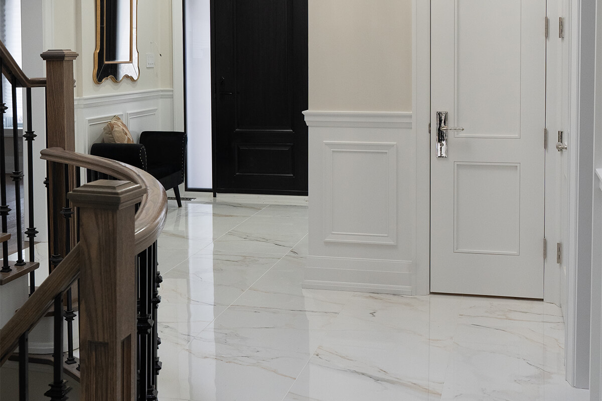 Lifestyle Construction A hallway with white marble floors, a wooden staircase featuring black railings, and a stylish black armchair against the wall showcases the elegance of Toronto Renovation. A sleek white door with a metallic handle complements the striking black front door in this expertly crafted space. Home Renovation In The GTA