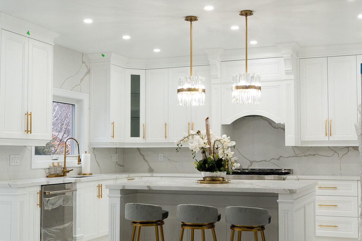 Lifestyle Construction A modern kitchen featuring white cabinets, marble countertops, and two pendant lights above an island. Three stools line the island, and a window is visible in the background. This stylish space is a prime example of Toronto Renovation expertise. Home Renovation In The GTA