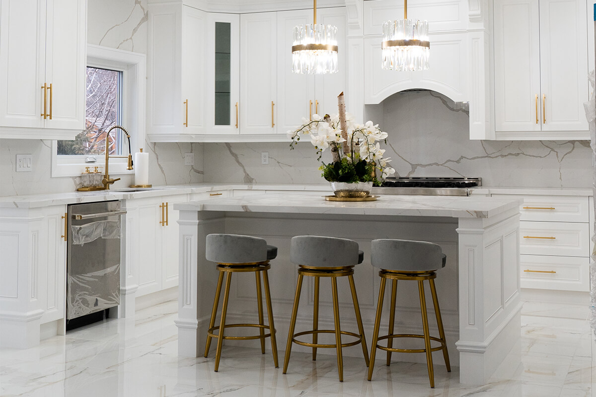 Lifestyle Construction Modern kitchen with white cabinetry, marble countertops, gold accents, and three gray barstools around a central island. Two pendant lights hang above the island, exemplifying the sleek design often seen in Home Renovation Vaughan projects. Home Renovation In The GTA