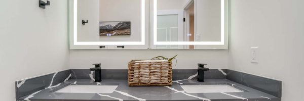 Lifestyle Construction A modern bathroom vanity with two illuminated mirrors, a gray and white marble countertop, double sinks, black fixtures, and a wicker basket with a plant in the center—perfect for your next Toronto Renovation. Home Renovation In The GTA